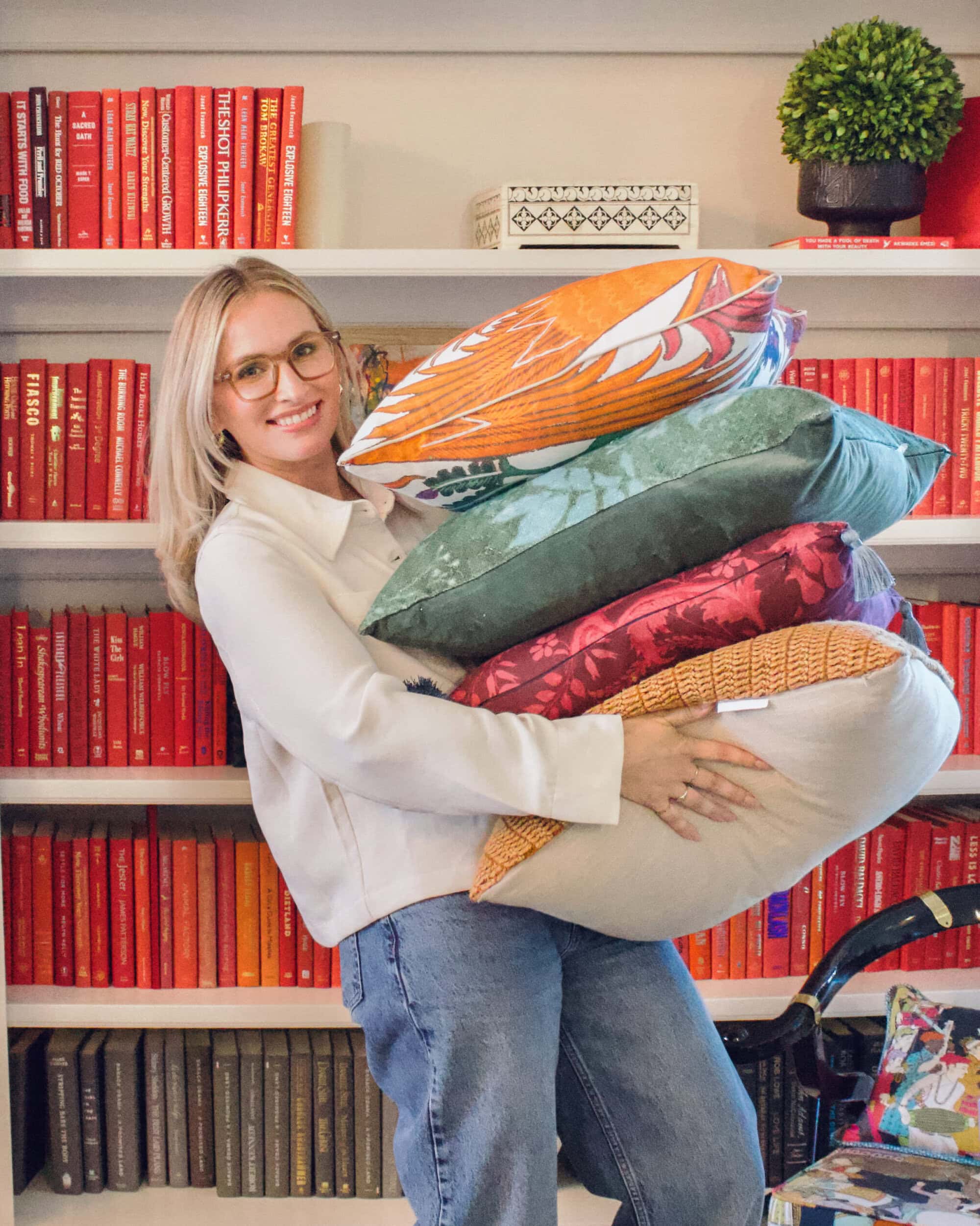 A friendly portrait of a female Crimson team member smiling and holding a stack of diverse, high-texture commercial pillows (velvet, orange-patterned, and damask). She is standing in front of a professionally styled bookshelf with color-coded red and black books for a luxury commercial project.