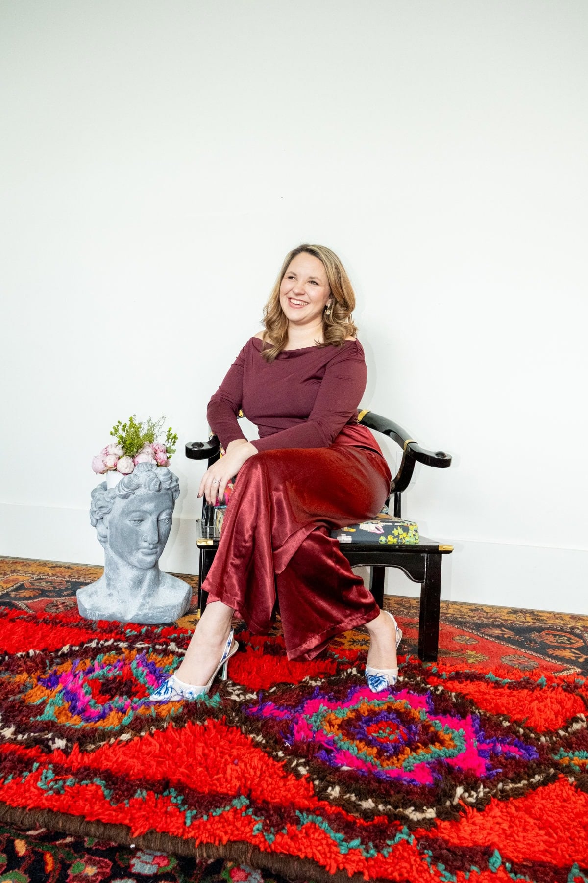 A woman in a maroon top and red velvet pants sits on a black chair, smiling, with a flower vase shaped like a head and a colorful red patterned rug beneath her.