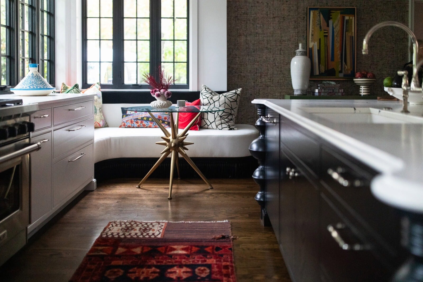 Modern kitchen with dark cabinets, white countertops, a patterned rug, a cushioned window seat with colorful pillows, and a small round table in front of the window.