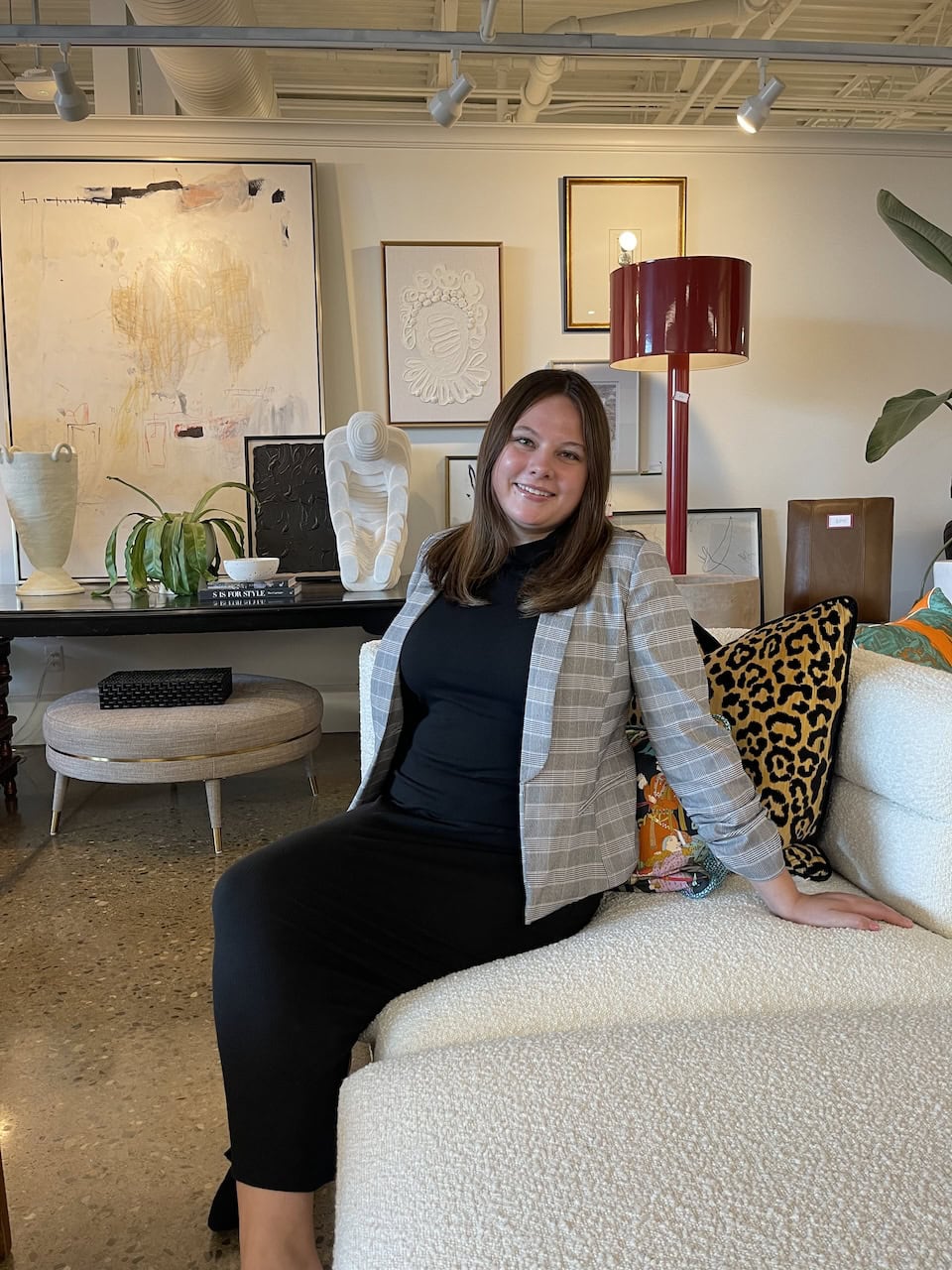 A woman in a plaid blazer sits on a white couch in a modern, art-filled living room with contemporary decor and wall art by Crimson Design Group.