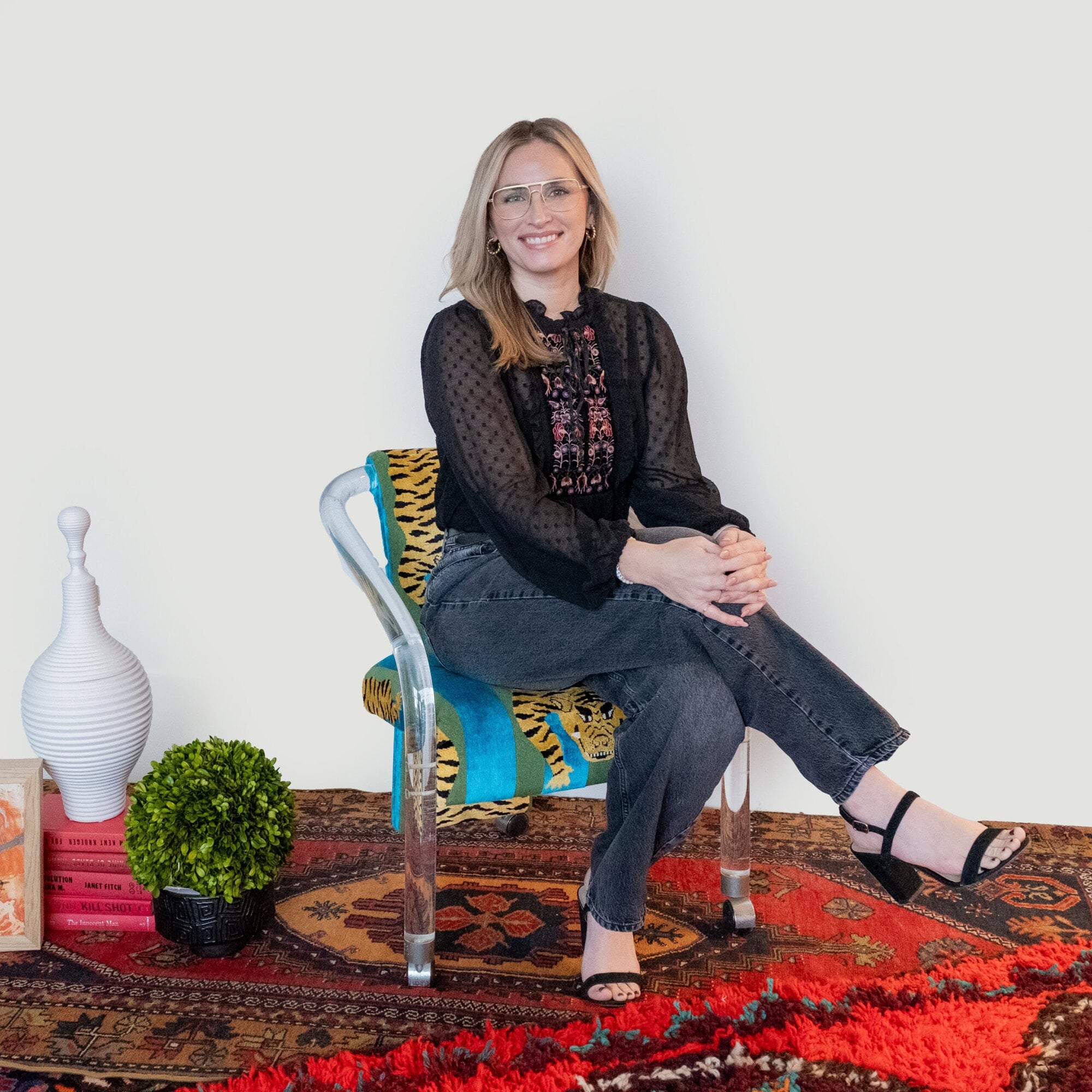 A woman with blonde hair and glasses sits in a patterned chair on a rug.
