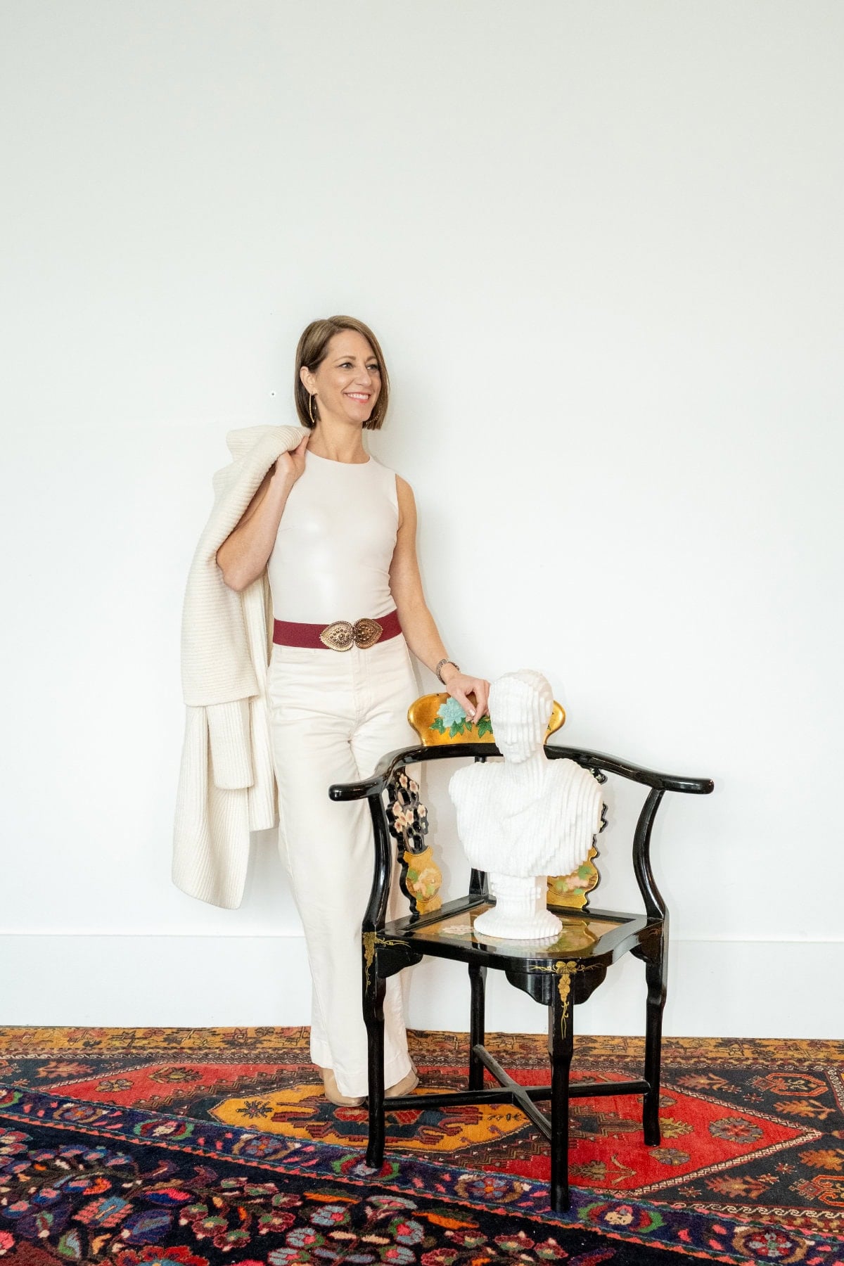 A woman in cream-colored clothing stands beside a black chair with a white bust on it, set against a white wall and patterned rug.