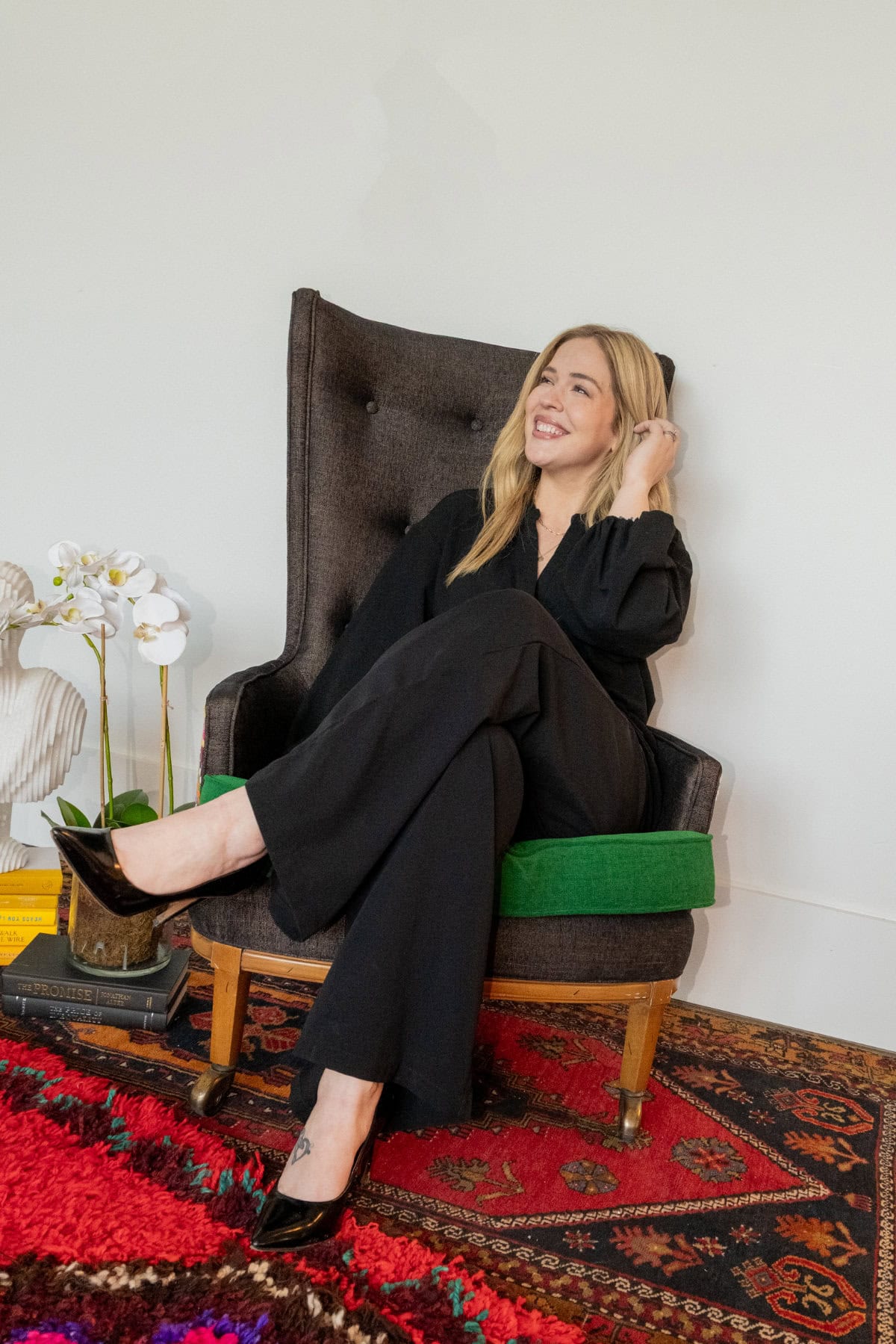 A woman in black clothing sits on a dark armchair with a green cushion, smiling and leaning back, beside a white orchid and stacks of books on a patterned rug.