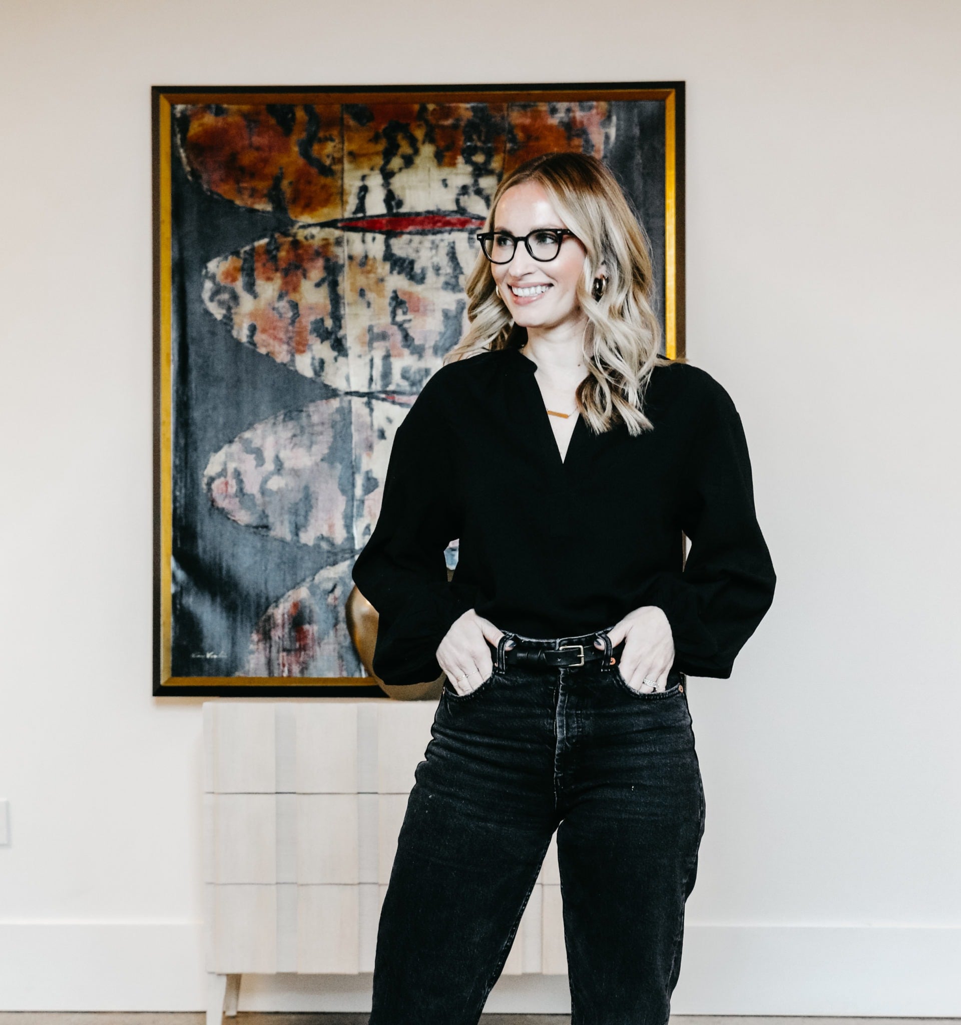 A person wearing glasses, a black top, and black jeans stands in front of abstract artwork and a light-colored dresser styled by Crimson Design Group.