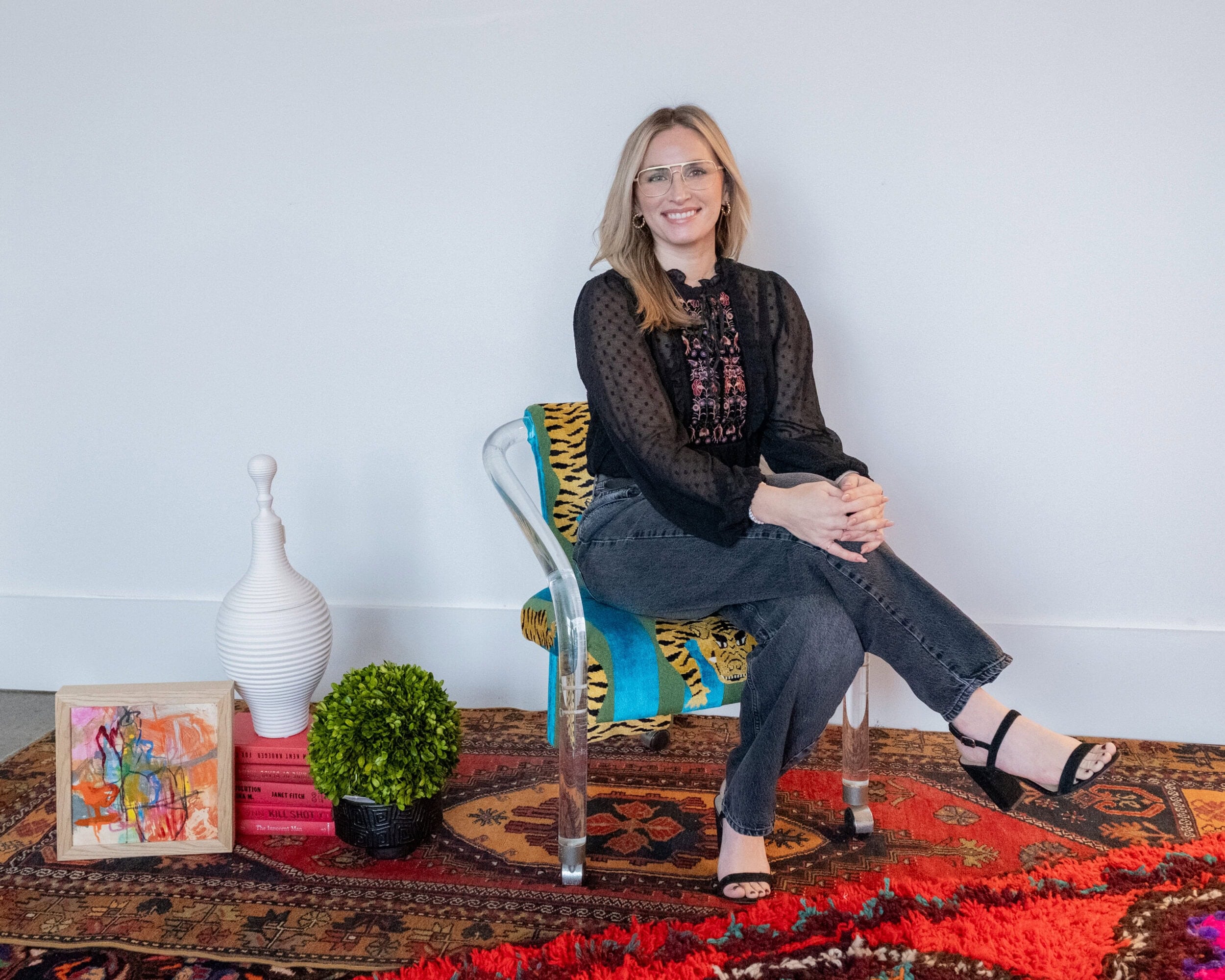 A woman sits on a clear chair with a patterned cover, legs crossed, surrounded by decorative items including books, a plant, a vase, and a small painting on a patterned rug.