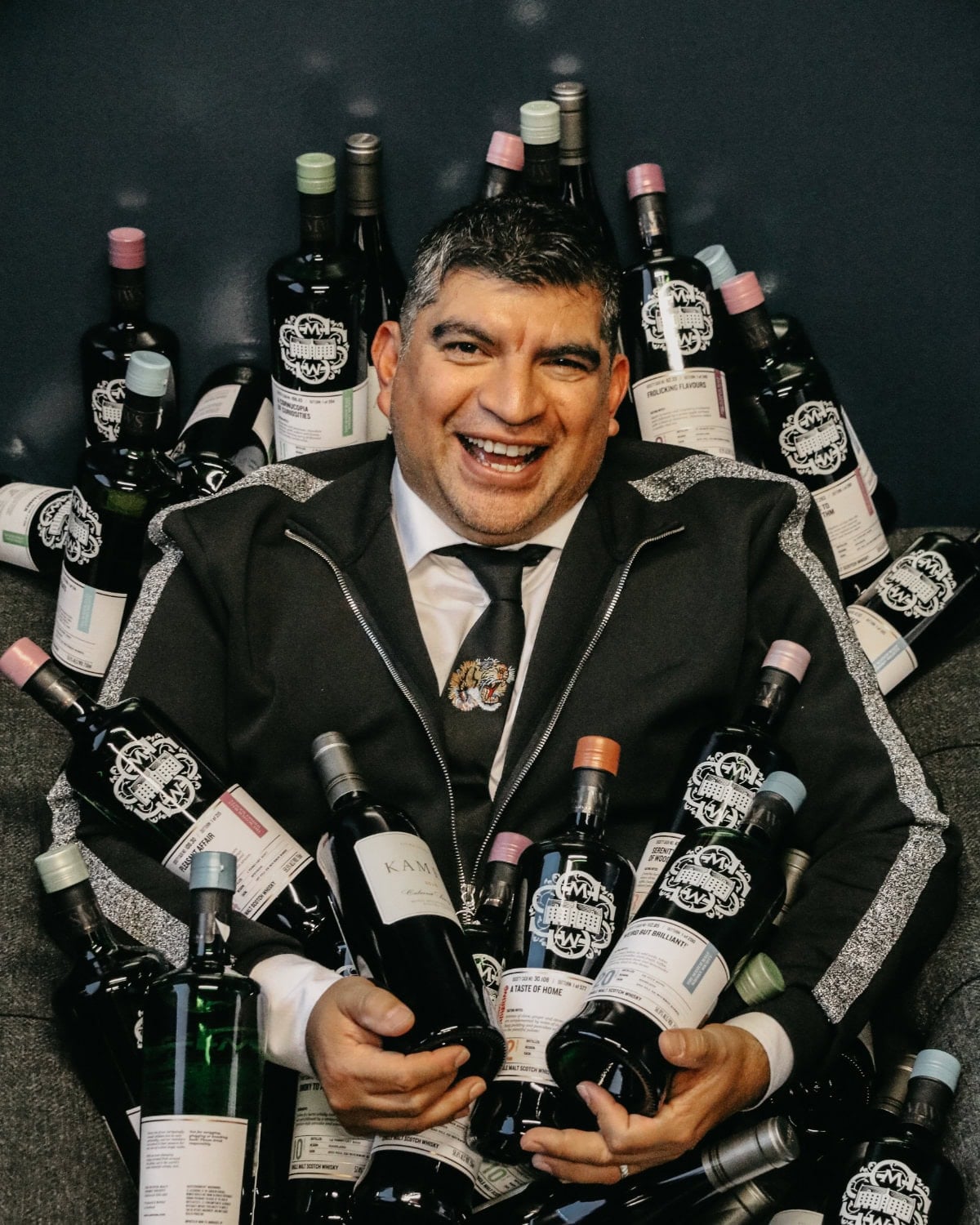 A man in a black suit sits smiling, surrounded by numerous bottles of wine with various labels against a dark background.