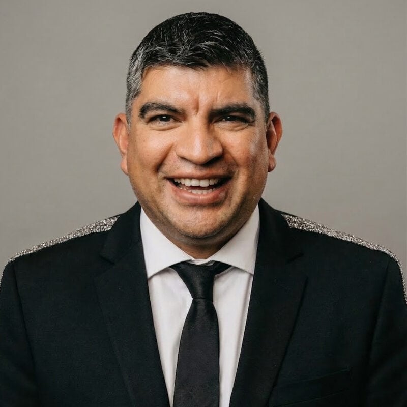 A man in a black suit, white shirt, and black tie is smiling in front of a plain grey background.