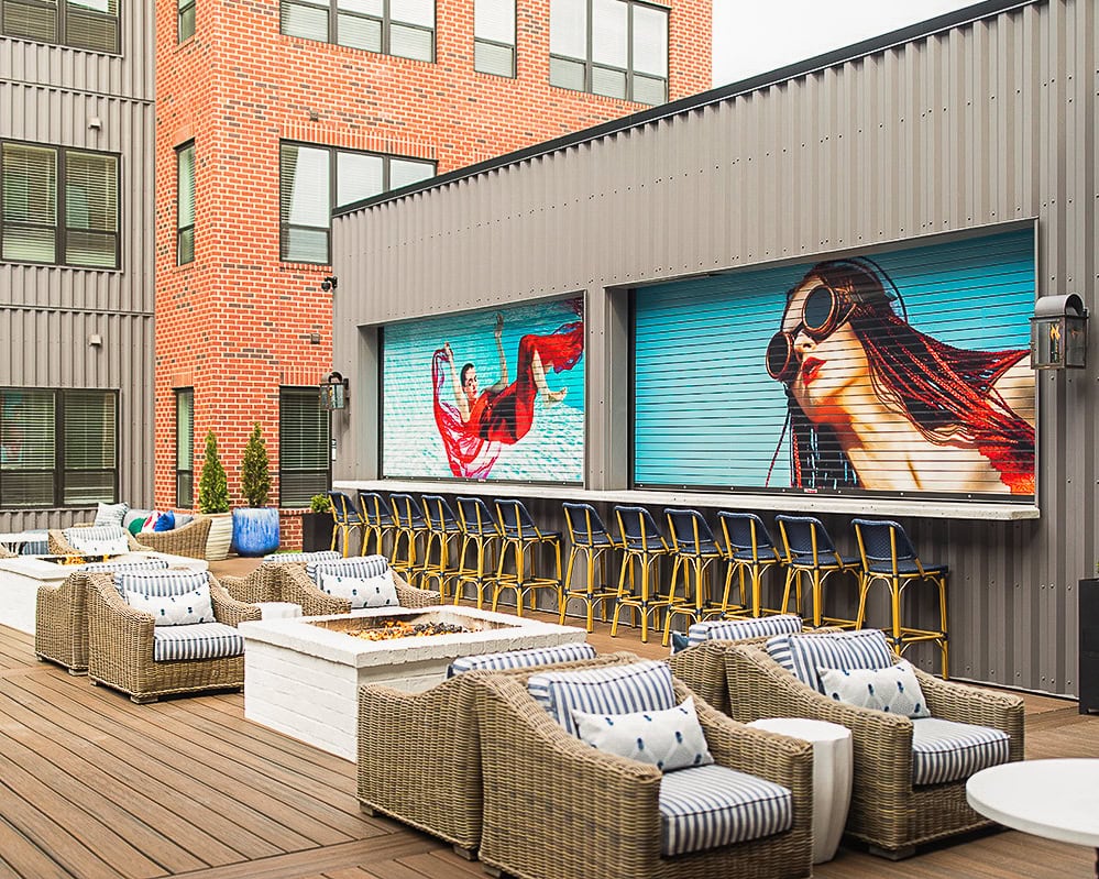 Outdoor patio area with wicker lounge chairs, a bar lined with stools, and colorful murals of women on a metal wall. Brick building with large windows in the background.