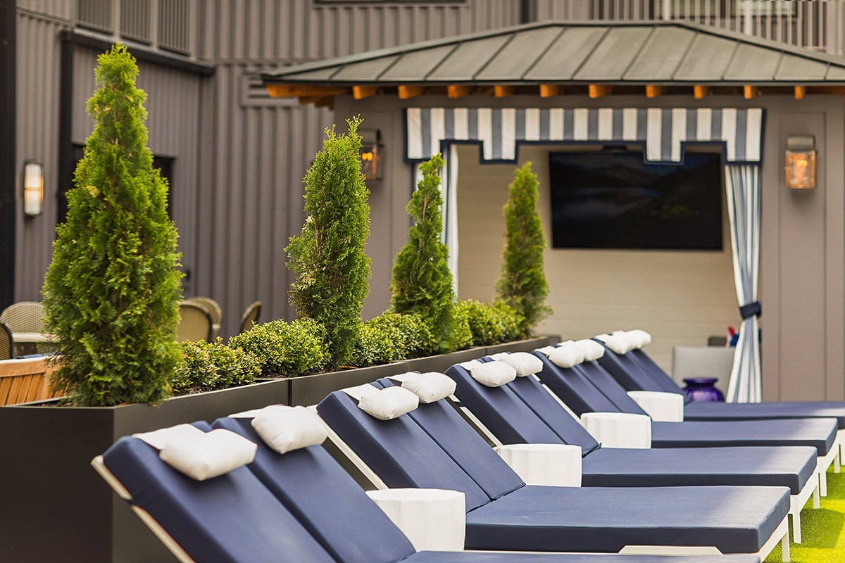 A row of navy blue lounge chairs with white headrests is arranged beside tall potted shrubs, in front of a cabana with striped blue and white curtains.