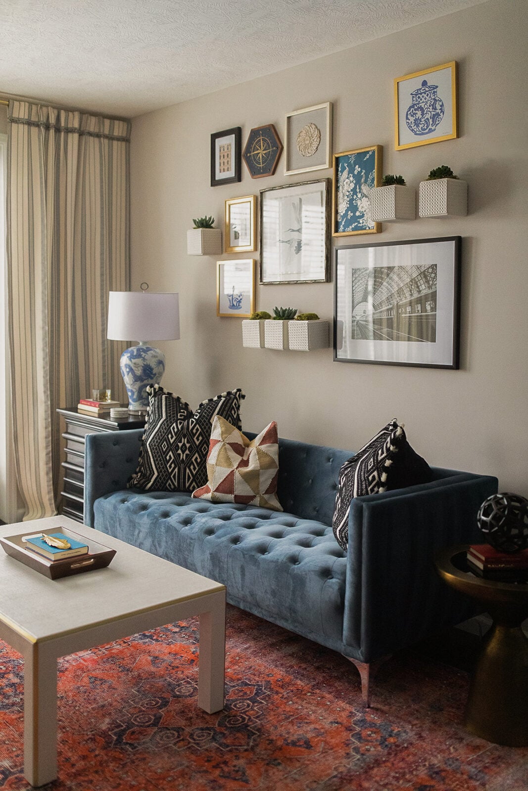 A modern living room with a blue tufted sofa, patterned pillows, a white coffee table, wall art, small plants on shelves, a lamp, and a red-patterned rug.