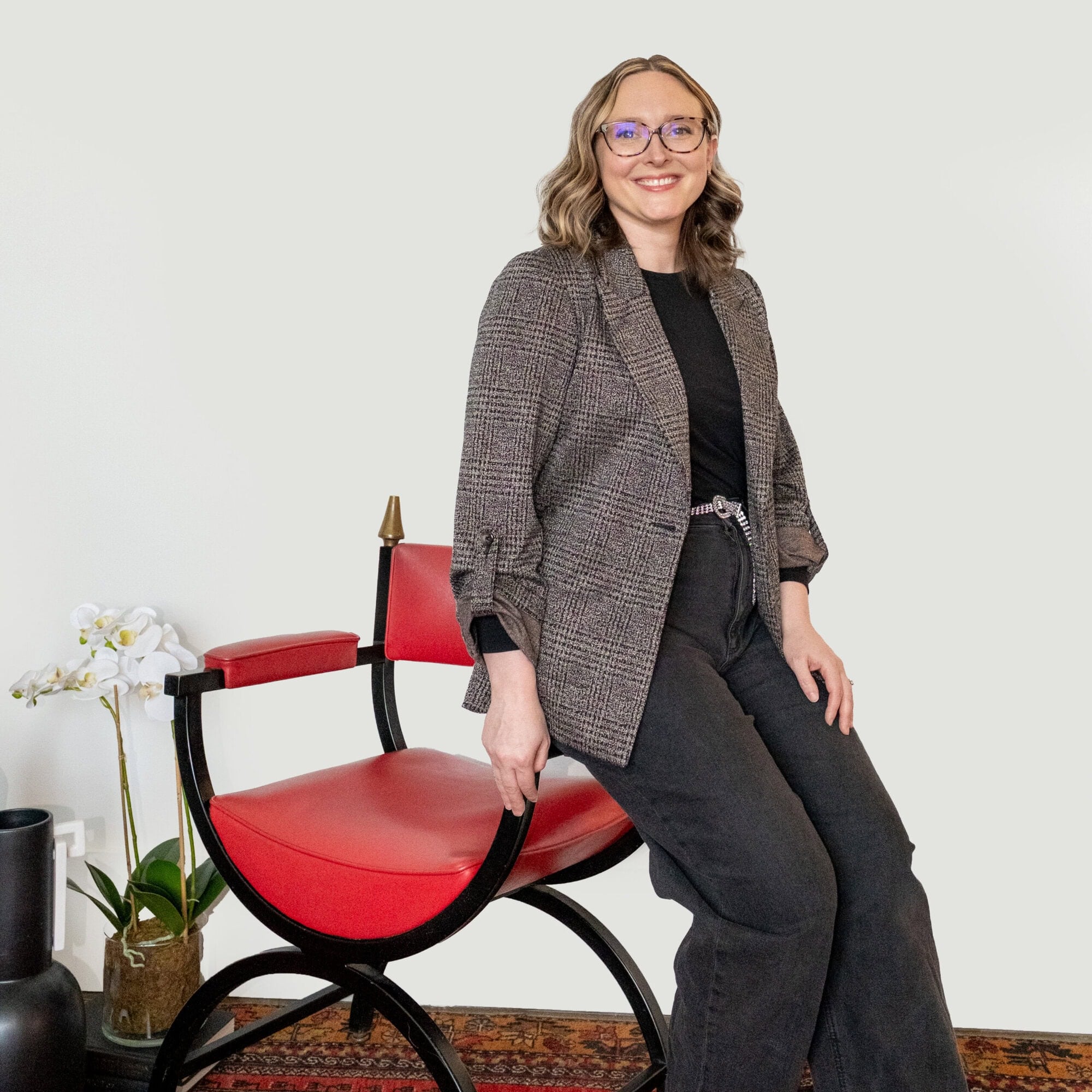 A woman in a blazer and glasses leans against a red and black chair.