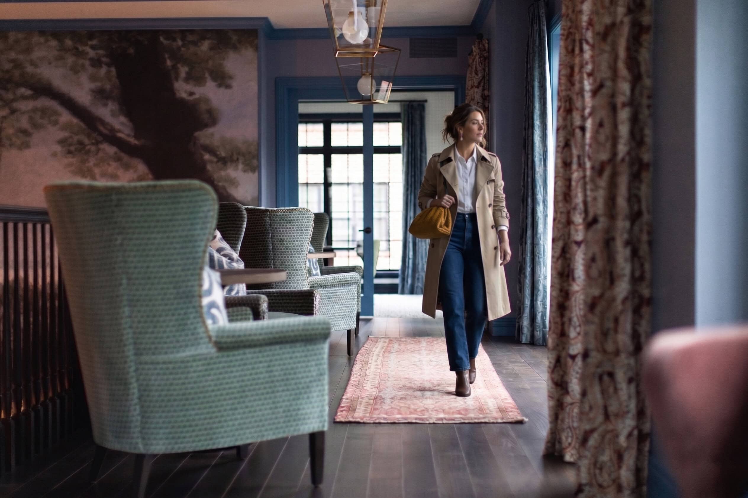 A woman wearing a trench coat and carrying a yellow bag walks through a stylish, softly lit lounge with patterned chairs and large windows.