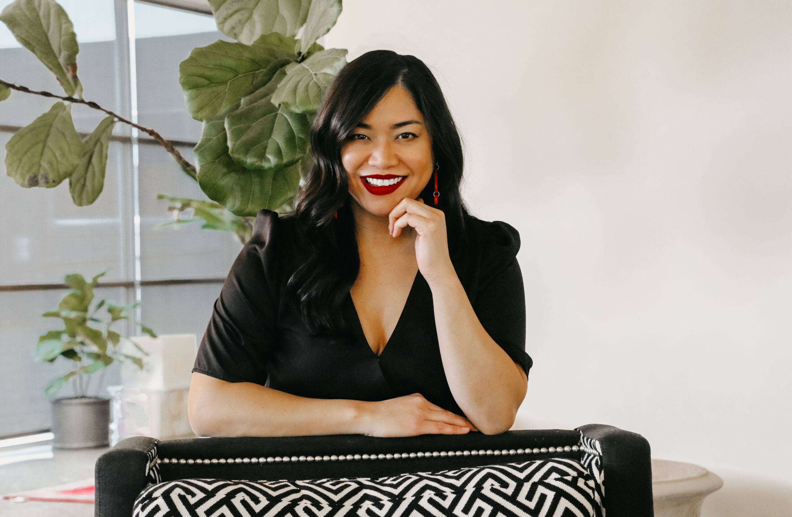 A woman with long black hair and red lipstick sits in a black and white chair, smiling at the camera. Styled by Crimson Design Group, the space features a large green plant in the background.