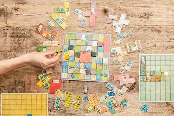 A person's hand moves a piece on a colorful patchwork-themed board game, designed with Crimson Design Group flair, as various game pieces and boards are spread out on a wooden table.