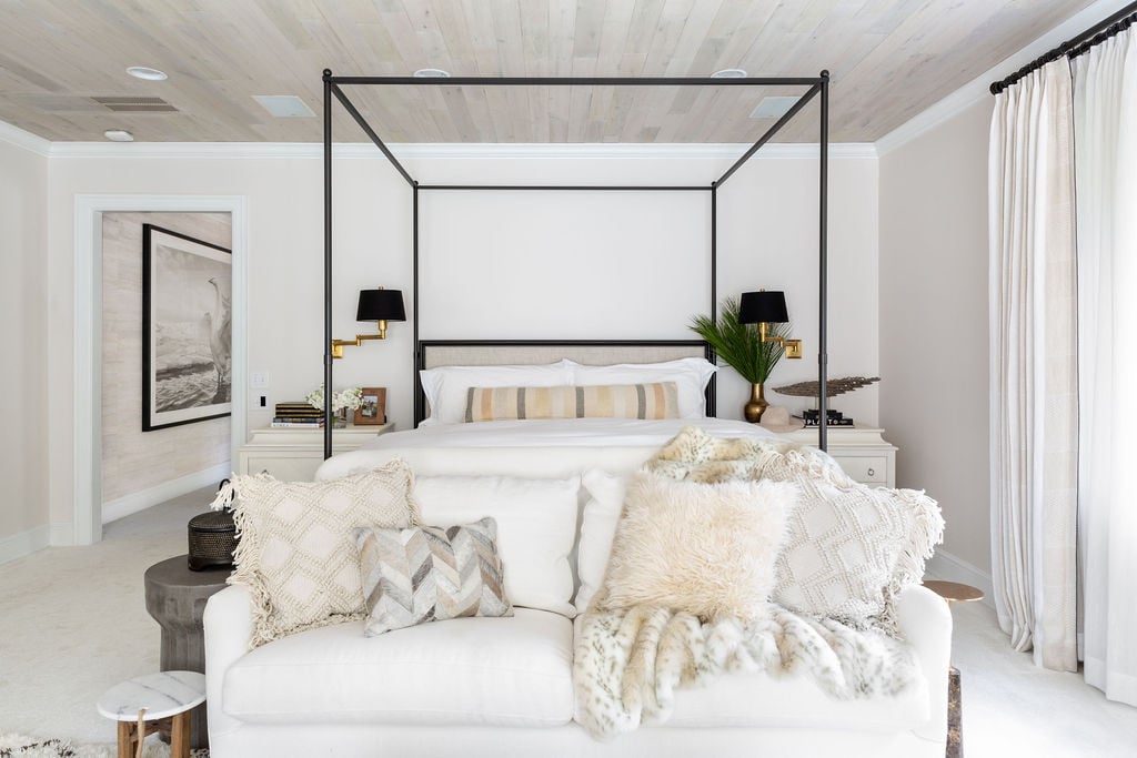 A modern bedroom with a four-poster bed, white bedding, neutral pillows, a white sofa at the foot, and beige walls with light wood accents. Large curtains cover a window on the right.