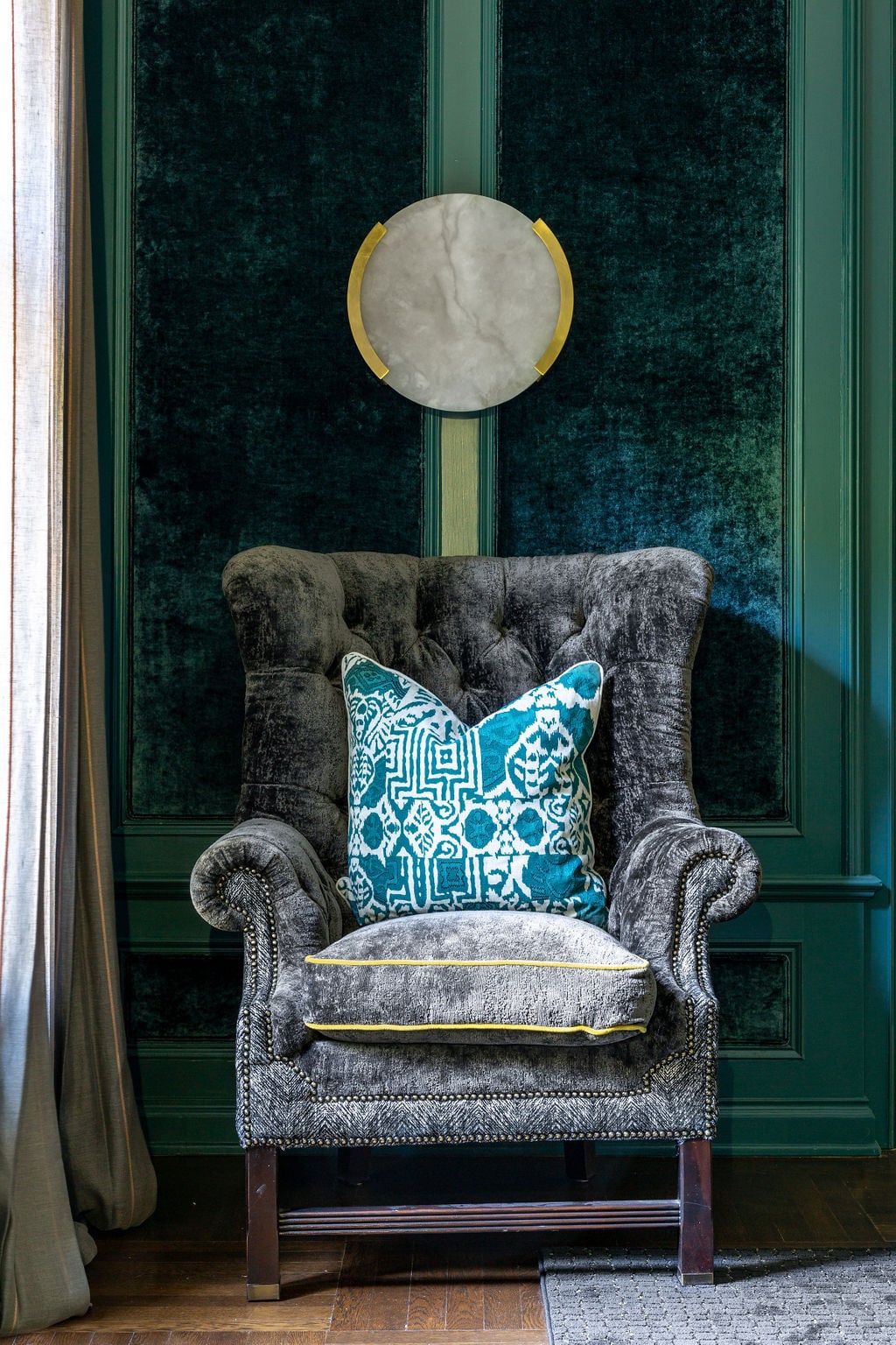 Gray velvet armchair with a blue patterned pillow against a dark green paneled wall. A round white wall decor hangs above the chair. Curtain and rug are partially visible.