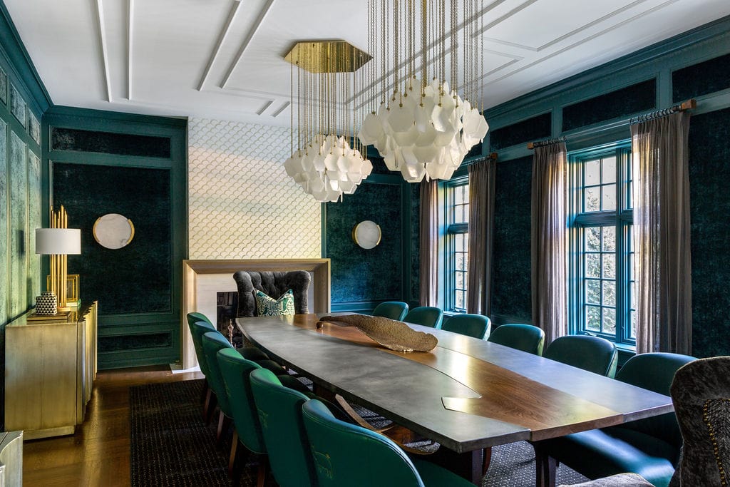 A modern dining room with a large wooden table, green upholstered chairs, geometric chandelier, dark walls, fireplace, and large windows with sheer curtains.