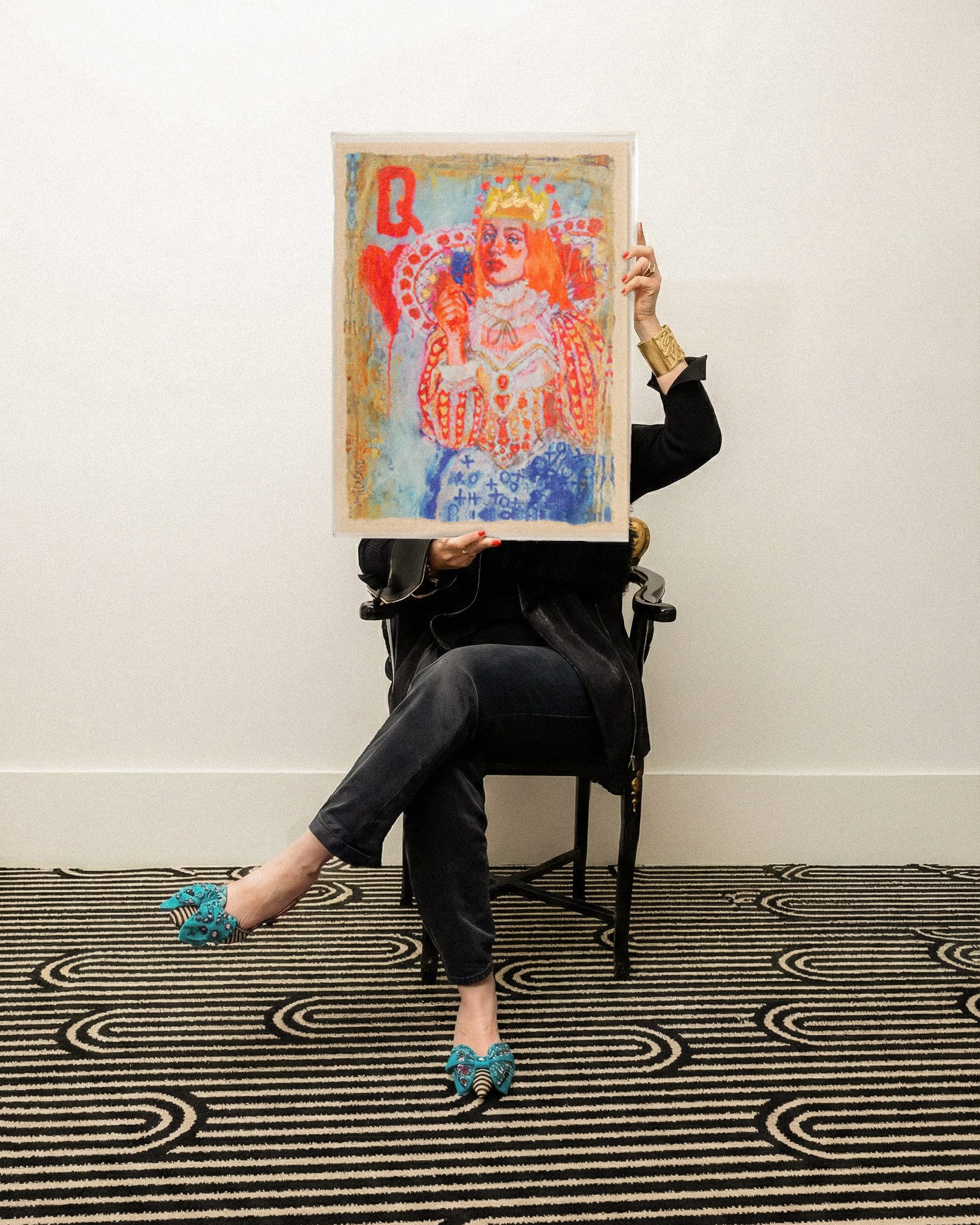 A person sitting on a black chair crosses their legs and holds a framed painting of a queen playing card, obscuring their face; the background is a white wall and patterned carpet.