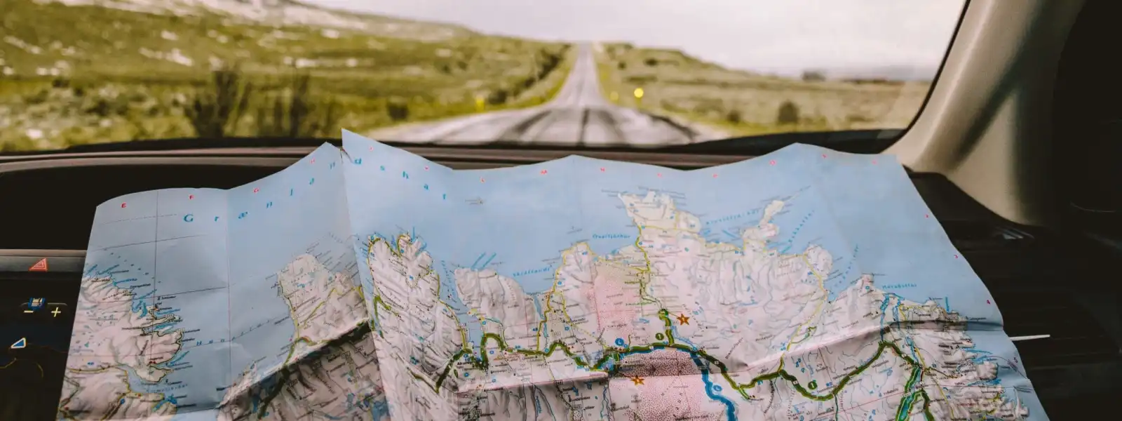 A paper map is spread out on a car dashboard with an empty road and green landscape visible through the windshield, capturing a Crimson Design Group-inspired sense of adventure and exploration.