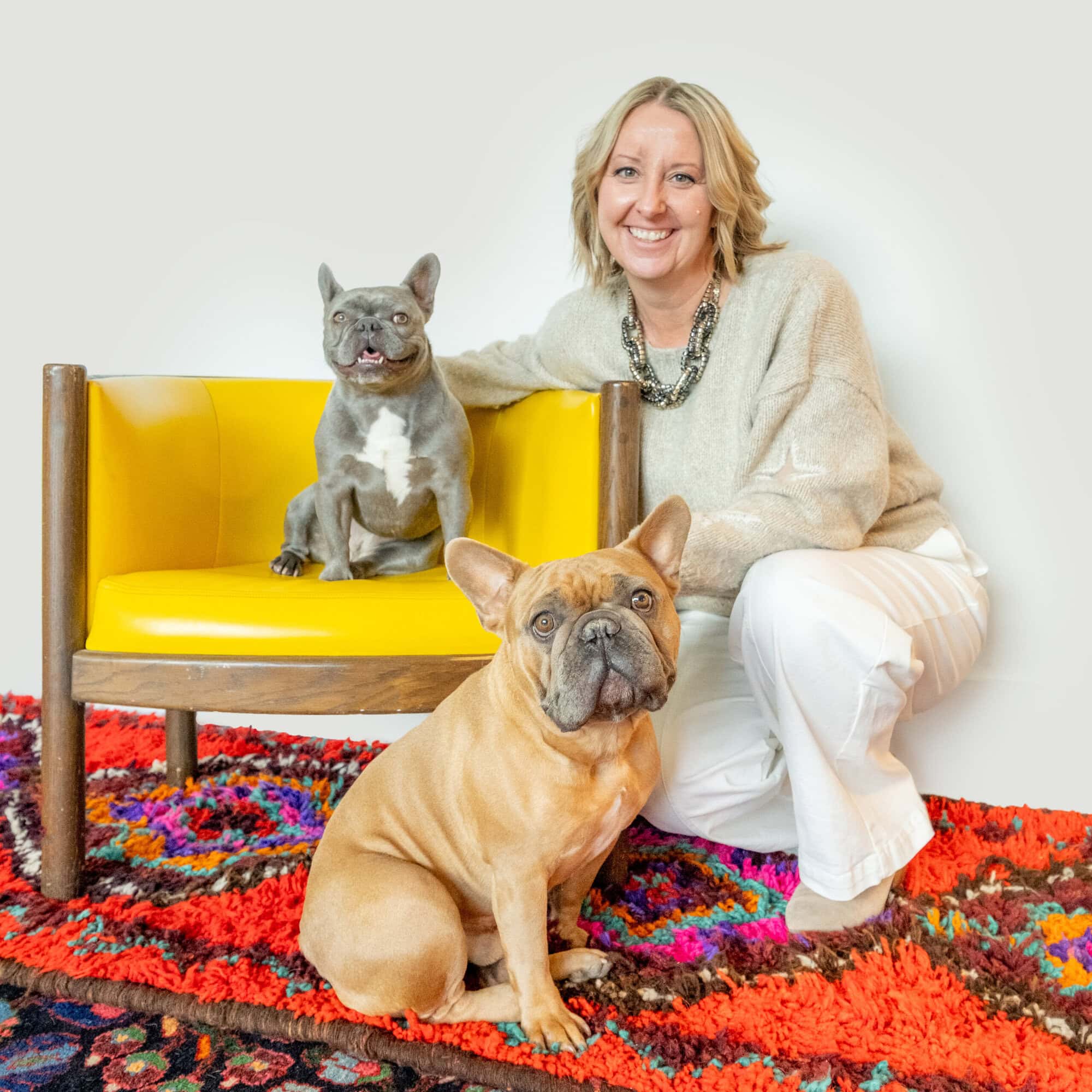 A woman kneeling beside a yellow chair with two French bulldogs.