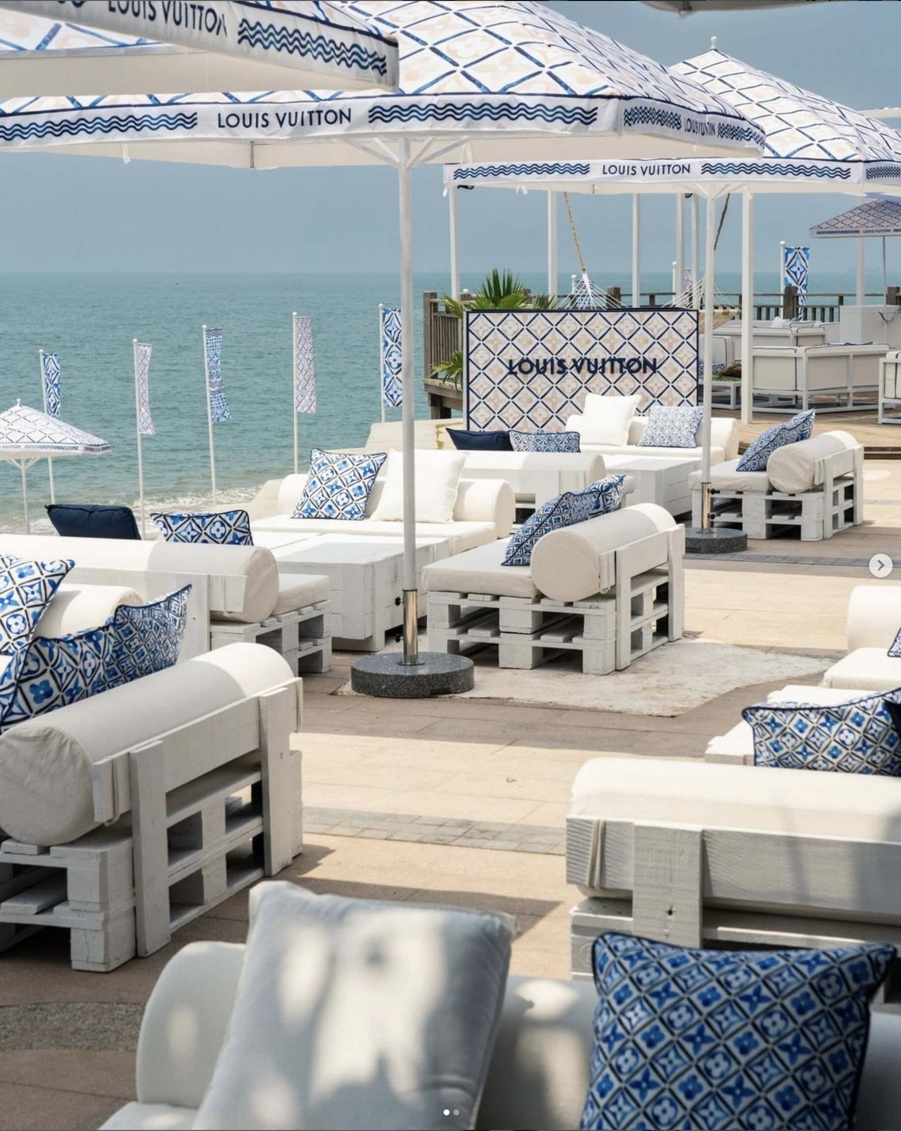 Outdoor lounge area by the sea with white wooden furniture, blue and white patterned cushions, Louis Vuitton branded umbrellas, and Crimson Design Group-inspired decor.