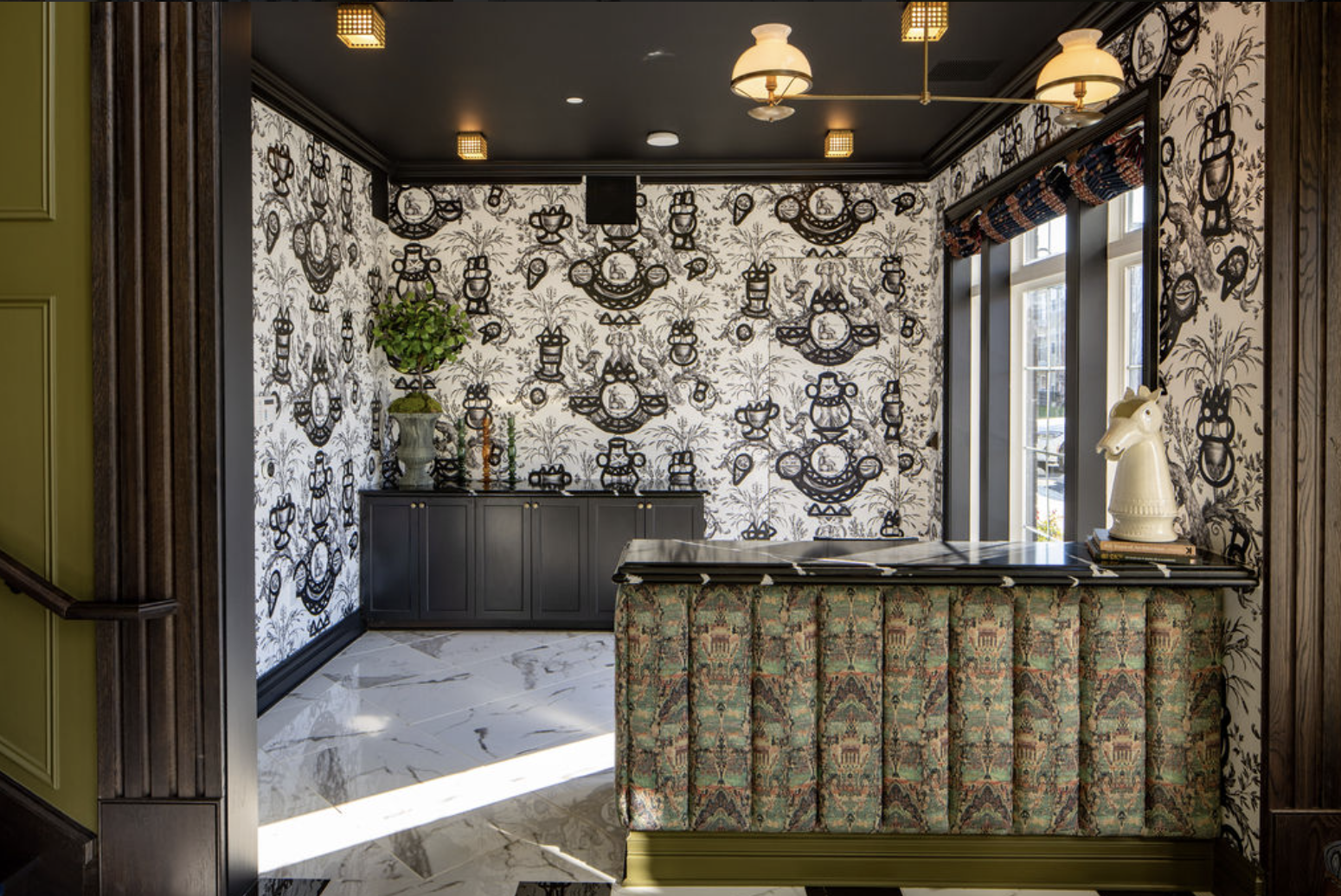 A patterned reception area by Crimson Design Group features a marble floor, black cabinets, black-and-white wallpaper, a floral arrangement, and a counter with a decorative horse head sculpture.