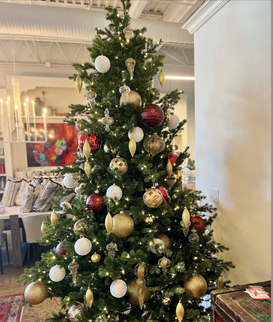 A decorated Christmas tree with gold, red, and white ornaments stands indoors near a dining area, showcasing colorful artwork in the background—a festive touch by Crimson Design Group.