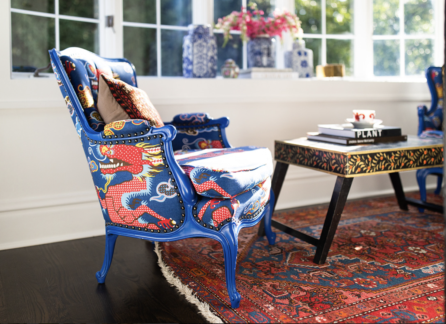 A blue upholstered armchair with a red and blue dragon pattern, styled by Crimson Design Group, sits beside a patterned coffee table on a red rug in a sunlit room with large windows and decorative vases.
