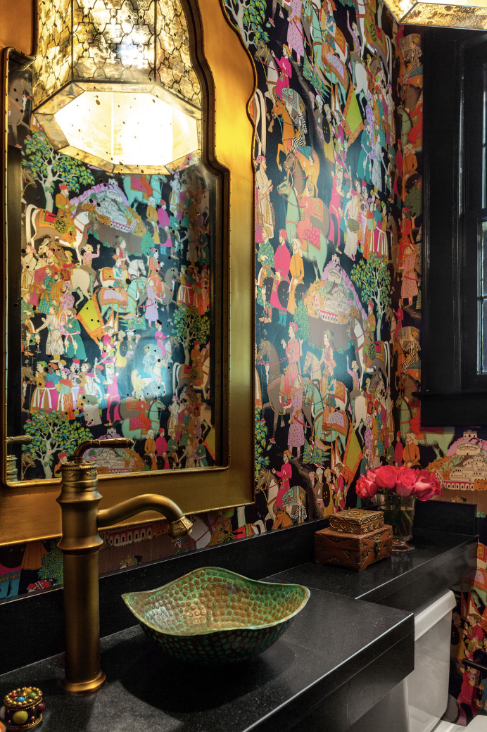 A bathroom designed by Crimson Design Group features colorful, ornate wallpaper, a gold-framed mirror, a green textured vessel sink on a black countertop, and a brass faucet. Pink flowers and decorative boxes add charm to the space.