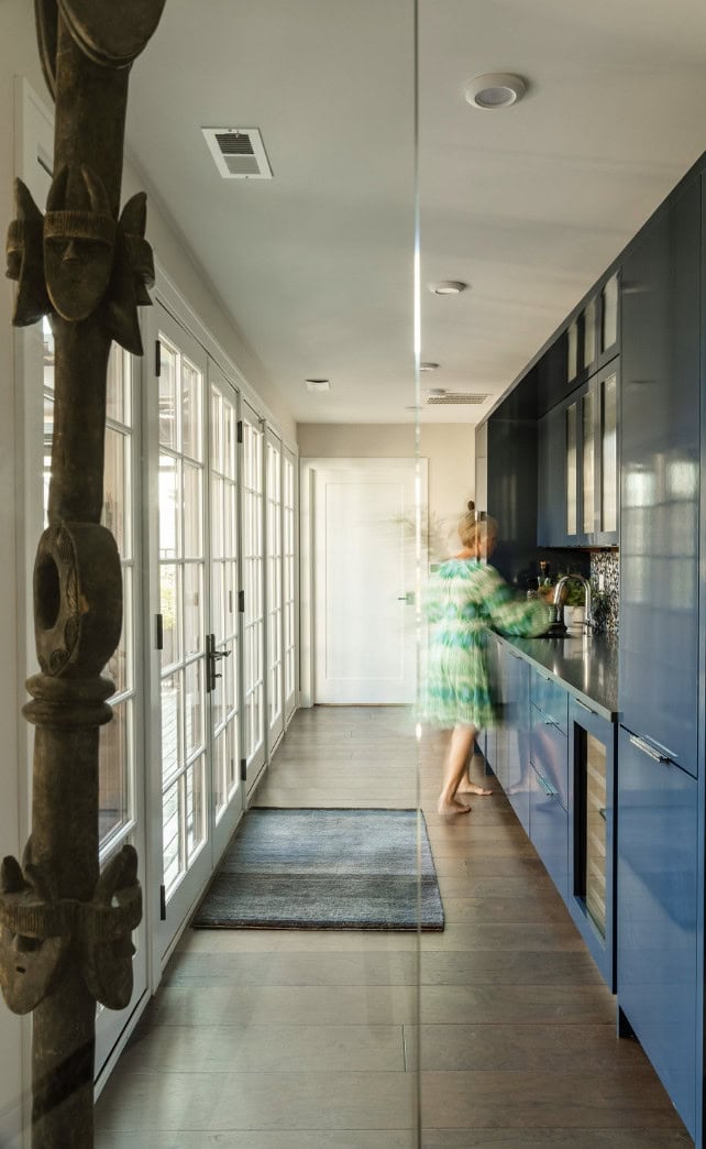 A hallway kitchen by Crimson Design Group features blue cabinets, wooden floors, and large windows; a blurred person stands at the counter preparing something.