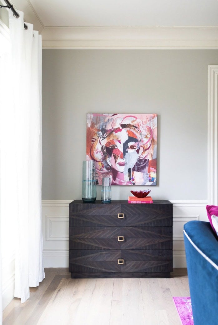A dark wood chest with three drawers sits against a pale gray wall, topped with glass vases, a red dish, and a pink book; abstract art hangs above in this Crimson Design Group inspired space.