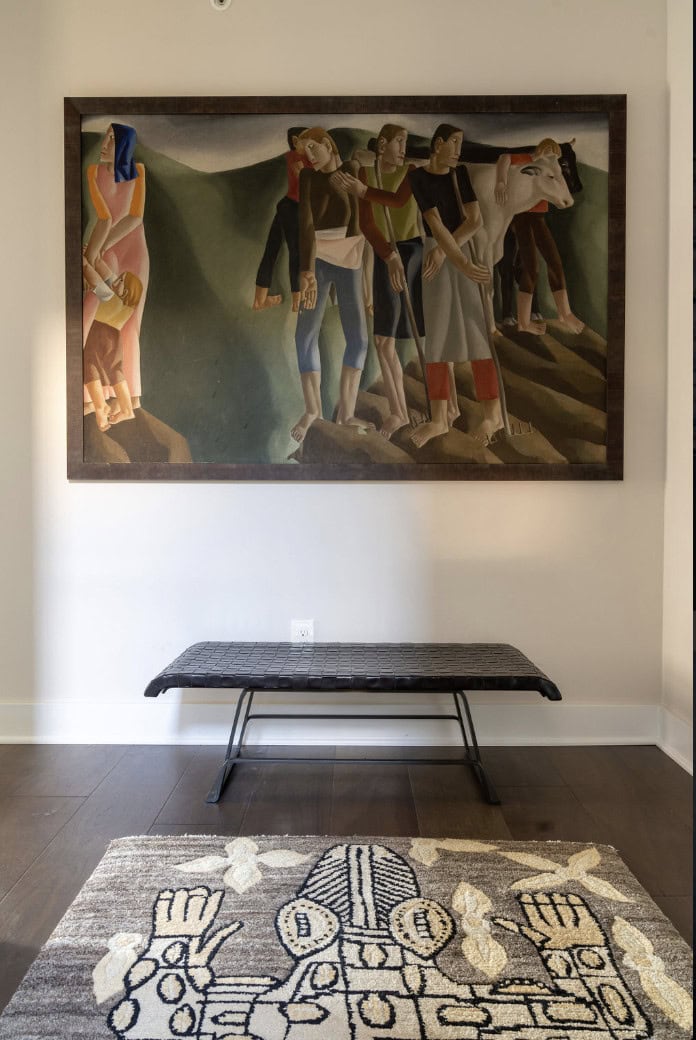 A large framed painting of abstract figures by Crimson Design Group hangs above a black bench; a patterned rug with abstract designs lies on the wooden floor below.