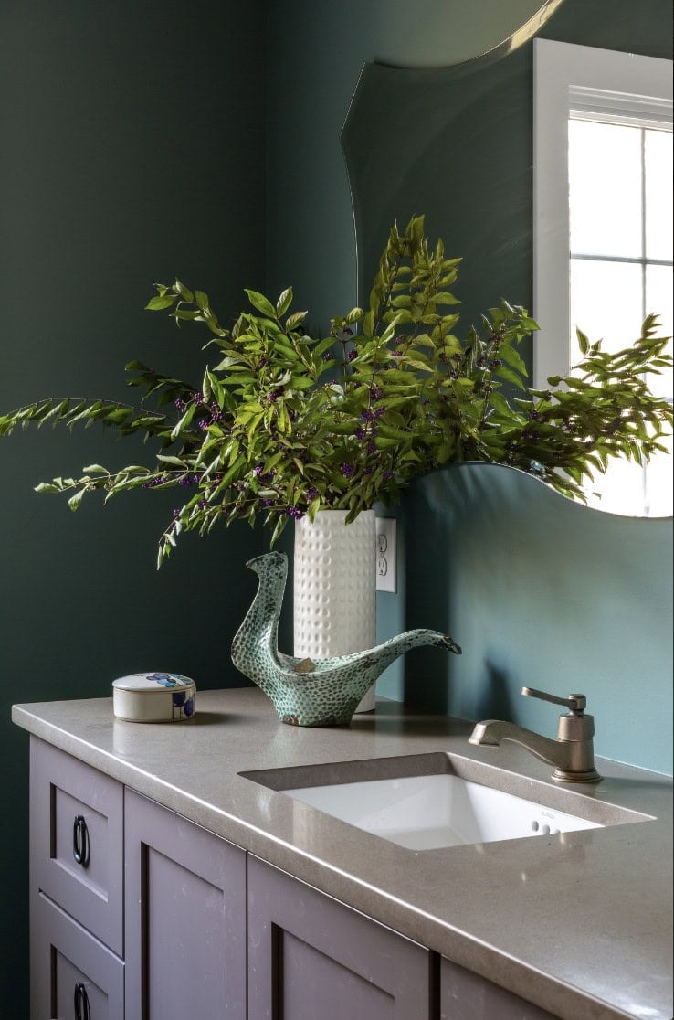 A modern bathroom by Crimson Design Group features a gray countertop, purple cabinets, a square sink, a decorative bird-shaped pitcher, a leafy vase arrangement, and a wall mirror next to a window.