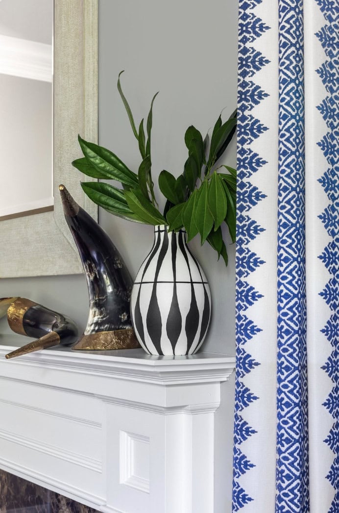 A white mantle styled by Crimson Design Group features a black-and-white striped vase with green leaves, two decorative horns, and a blue-patterned curtain in the background.