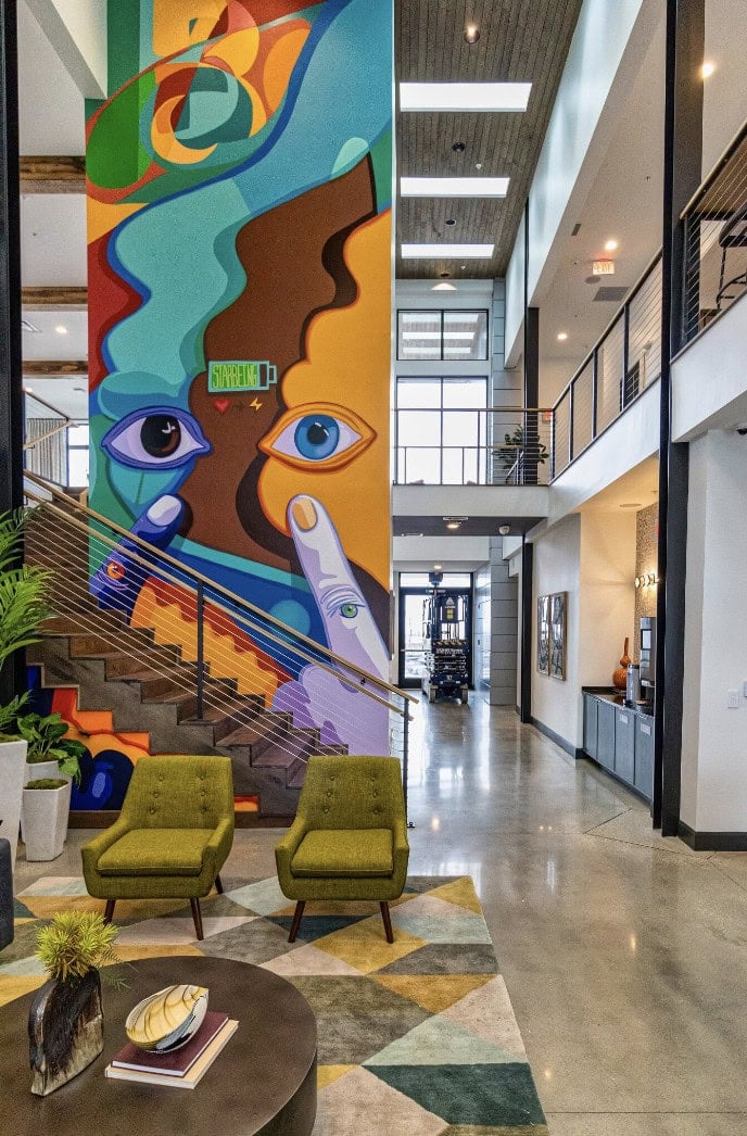 A modern lobby by Crimson Design Group with two green chairs, a geometric rug, and a colorful abstract mural featuring faces and hands beside a staircase.