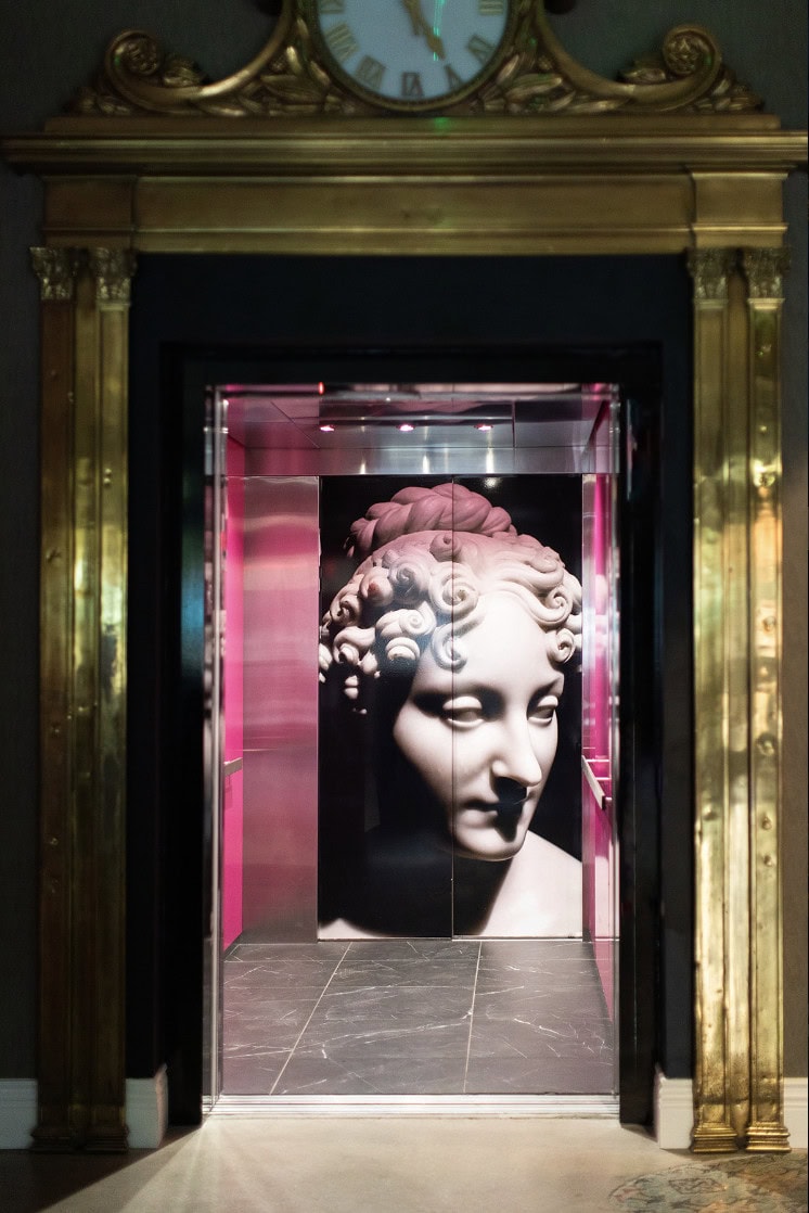 A marble statue head is depicted on the back wall of an elevator with pink side panels, framed by a gold ornate doorway and a clock above—a striking interior by Crimson Design Group.