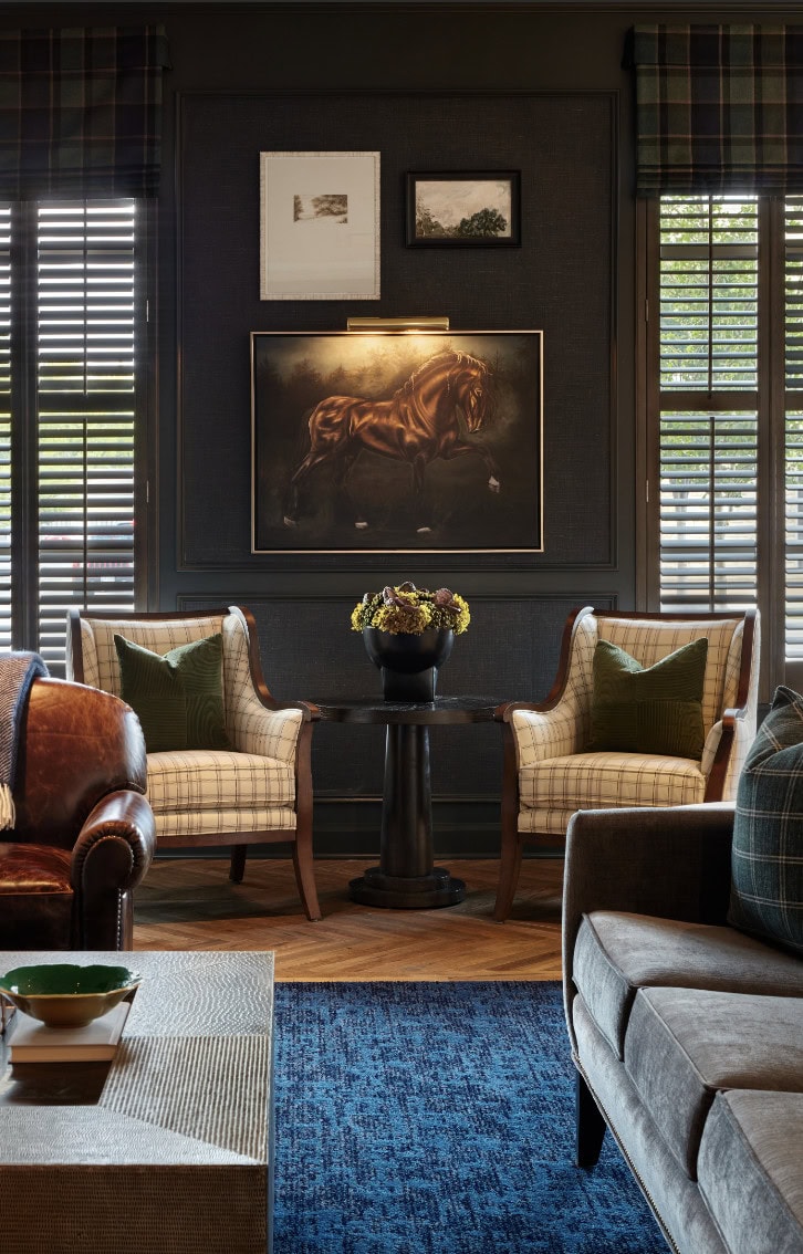 A sophisticated living room by Crimson Design Group features two plaid armchairs, a small round table with a flower arrangement, horse artwork on the wall, and large windows with wooden shutters.