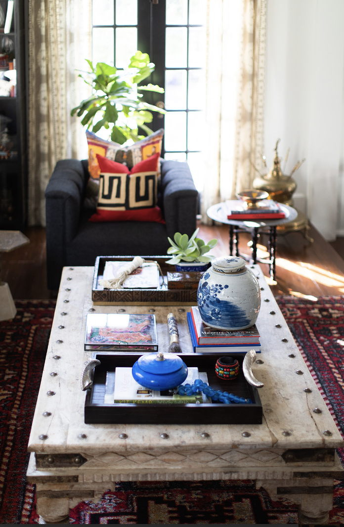 A living room by Crimson Design Group features a black armchair, red and gold pillows, a white coffee table with decorative items, books, a blue vase, and a plant by the window framed with sheer curtains.