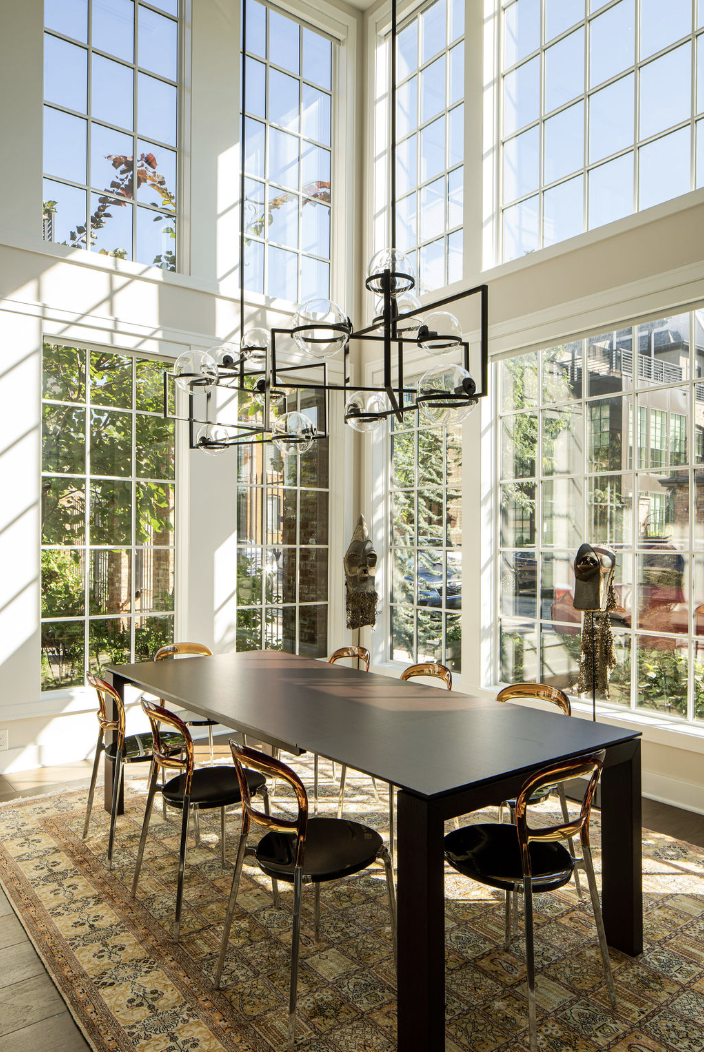 A modern dining room by Crimson Design Group features a black rectangular table, eight chairs, large windows, a patterned rug, and a geometric chandelier. Sunlight fills the space.