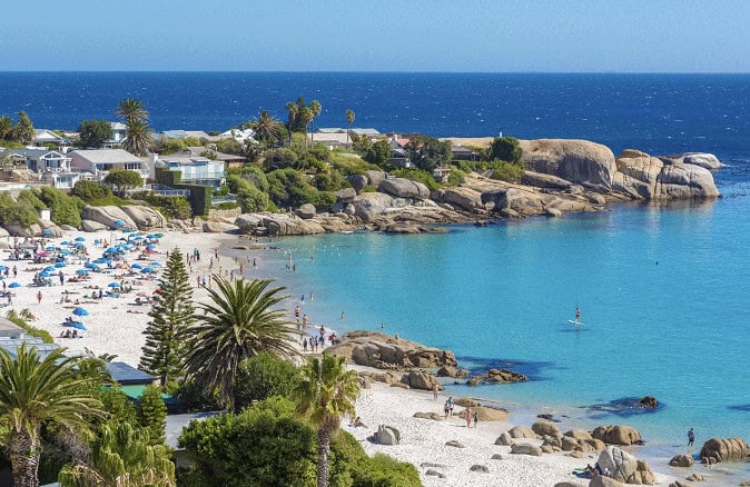 A beach with clear turquoise water, white sand, palm trees, and scattered sunbathers; houses and large boulders line the shore under a bright blue sky, echoing the vibrant aesthetic of Crimson Design Group.