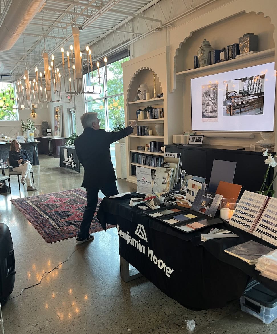 A person gestures toward a presentation screen in a modern, well-lit room with Benjamin Moore paint samples and Crimson Design Group color swatches displayed on a table in the foreground.