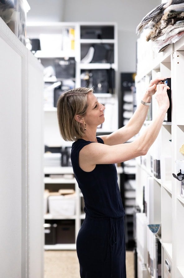 A woman in a sleeveless black outfit organizes items on white shelves in a neatly arranged storage room, reflecting the sophisticated touch of Crimson Design Group.