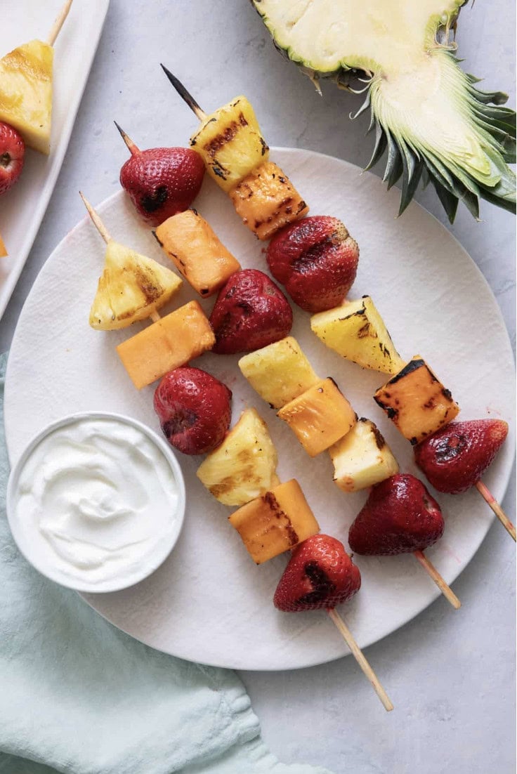 Three skewers with grilled strawberries, pineapple, and cantaloupe on a white plate next to a small bowl of yogurt and a slice of pineapple, styled in true Crimson Design Group fashion.