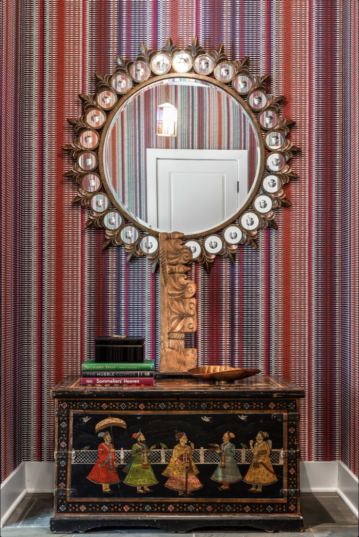 Round mirror with decorative sunburst frame above a chest featuring painted figures, set against red-striped wallpaper; books and a sculptural lamp accent the chest—an eclectic touch by Crimson Design Group.