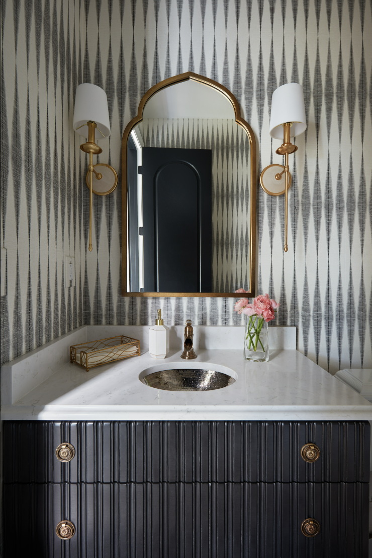 A bathroom vanity by Crimson Design Group features a marble countertop, gold fixtures, an arched mirror, two wall sconces, a vase of pink flowers, and geometric wallpaper.