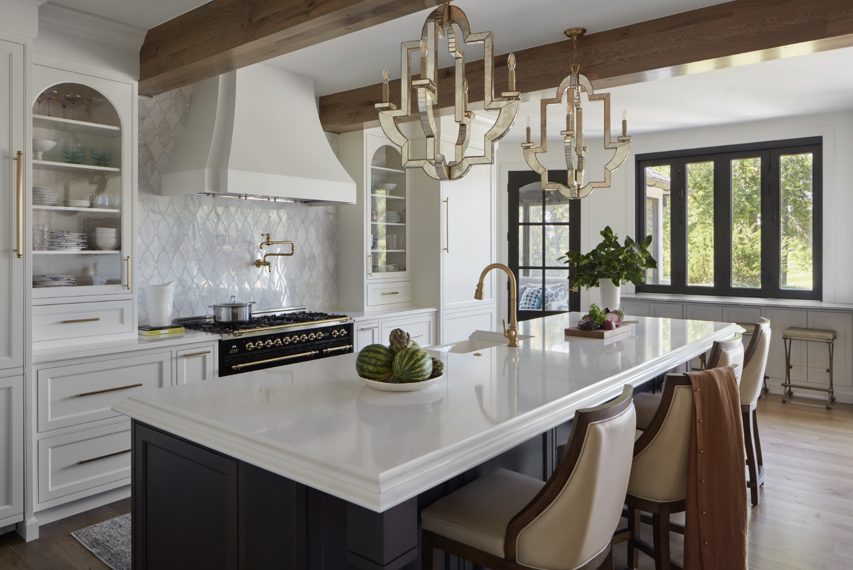 A modern kitchen by Crimson Design Group features a large white island, four chairs, gold fixtures, a black stove, white cabinetry, and two geometric pendant lights. Fruit and plants add fresh accents to the counter.