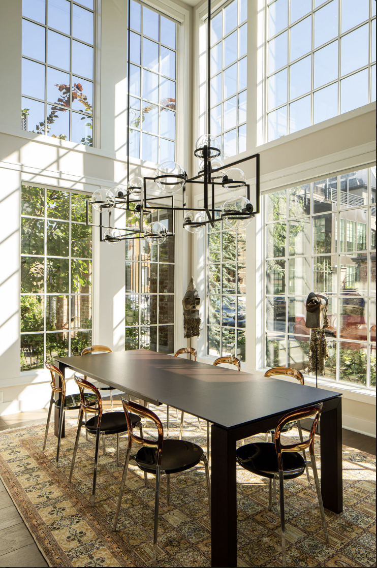 A modern dining room by Crimson Design Group features large grid windows, a black rectangular table, eight chairs, a geometric chandelier, and a patterned rug, all beautifully filled with natural sunlight.