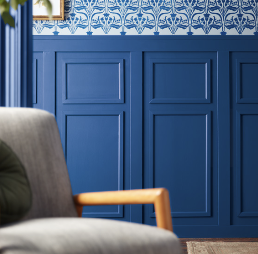 A close-up of a living room corner designed by Crimson Design Group, with a gray sofa featuring a wooden armrest, blue paneled walls, and patterned blue-and-white wallpaper above.