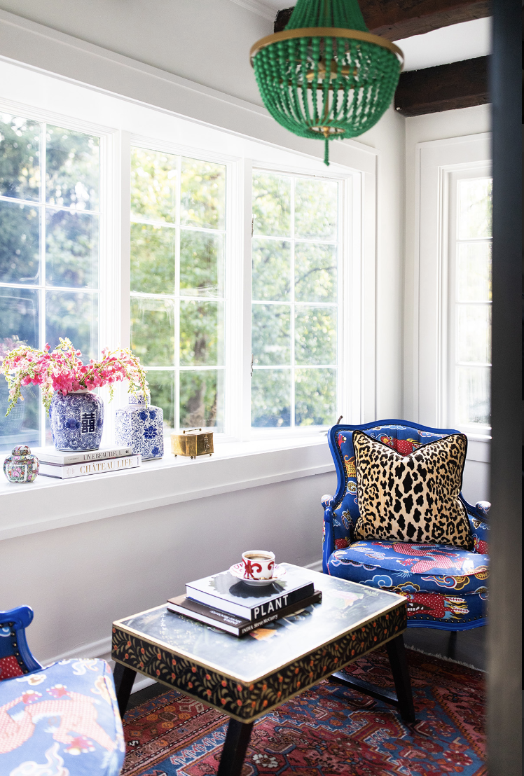 A bright sitting area by Crimson Design Group features blue patterned armchairs, a coffee table with books and a mug, a green pendant light, and large windows overlooking lush greenery.