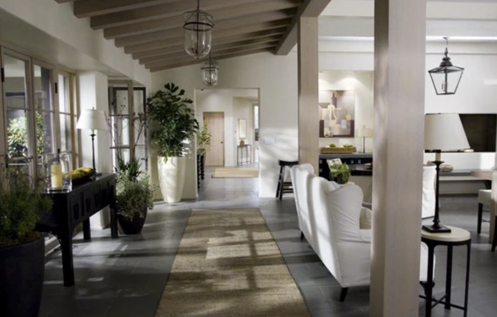 A modern living room by Crimson Design Group features white walls, exposed wooden beams, indoor plants, white sofas, a jute rug, and large windows flooding the space with natural light.